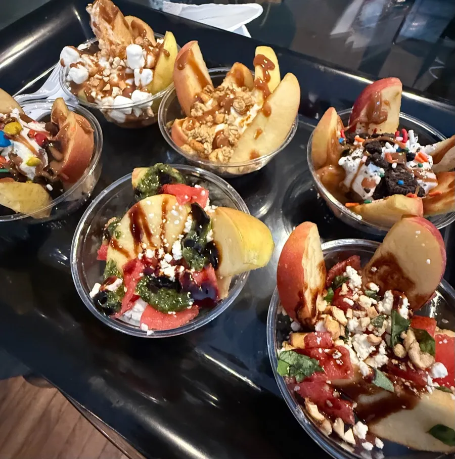 Six cups of sliced apples on a serving tray, each finished with different caramel, candy, granola and fruit toppings